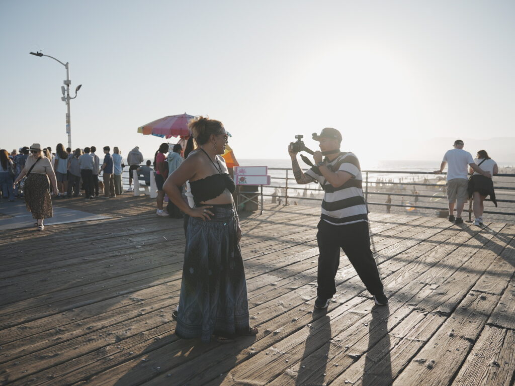 Photographer aiming camera at a woman posing on the wooden deck of Santa Monica Pier with the sun low over the Pacific