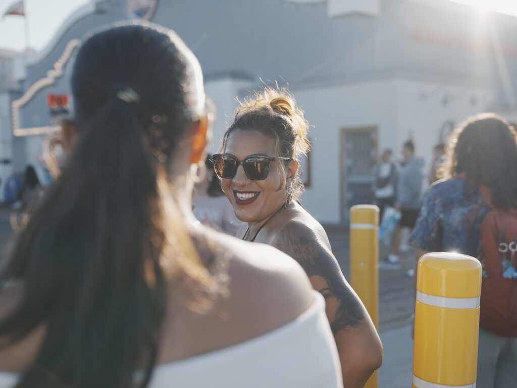 Cinematic close-up of a smiling woman in sunglasses on Santa Monica Pier, captured in warm afternoon light with soft focus foreground.