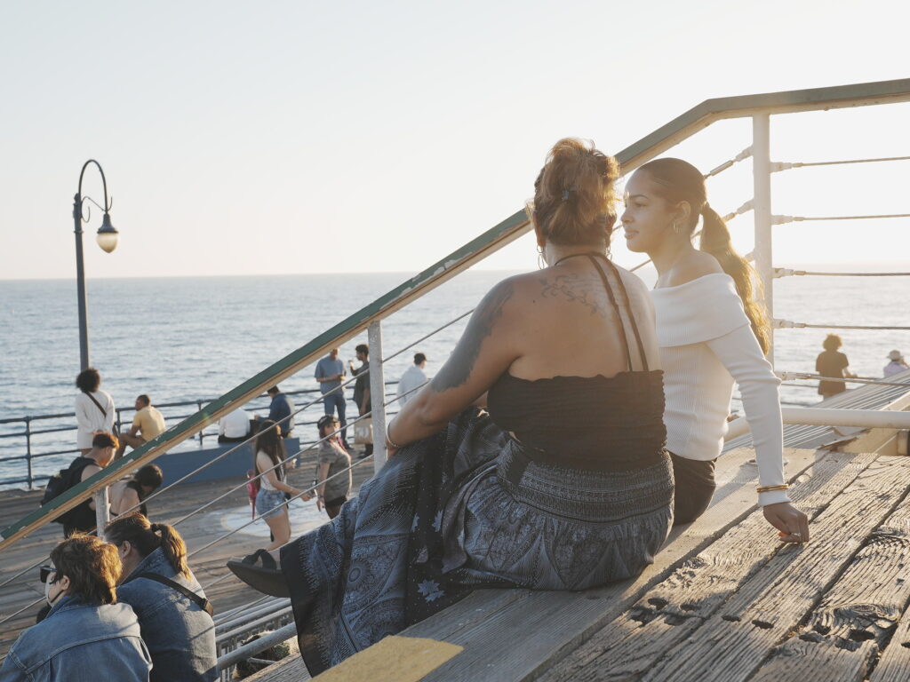 Mother and daughter sitting on wooden steps at Santa Monica Pier, talking as the sun sets over the Pacific Ocean.