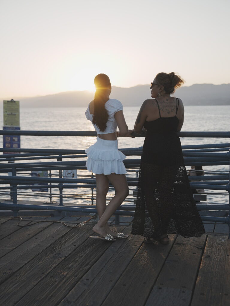 Mother and daughter standing at the railing on Santa Monica Pier, watching the sun set over the Pacific Ocean.