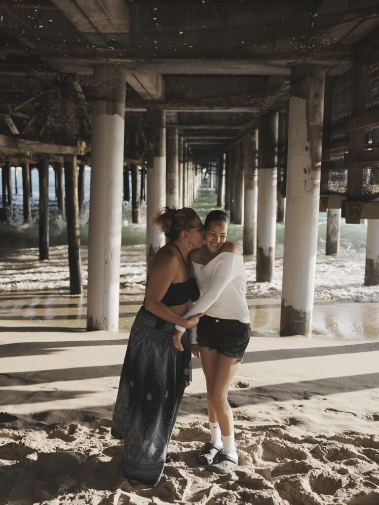 Mother kissing her daughter’s cheek under the Santa Monica Pier, standing on soft sand with sunlight streaming through the pillars in a safe, well-maintained setting.