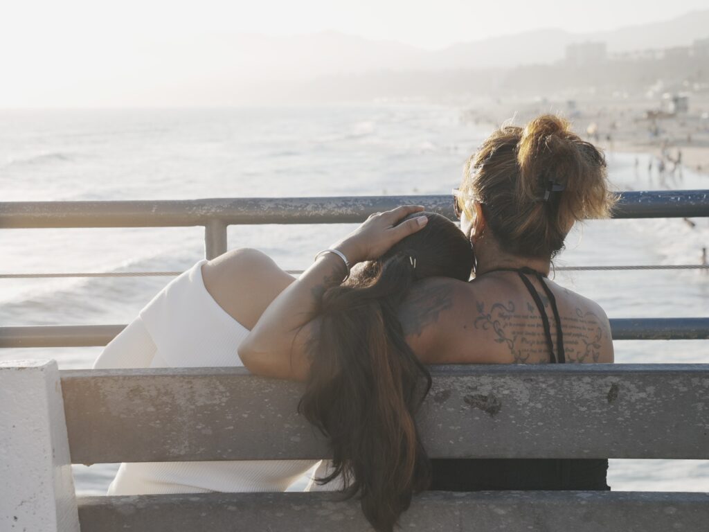 Mother and daughter sitting on a bench at Santa Monica Pier, with the daughter resting her head on her mother’s shoulder as they look out over the ocean.