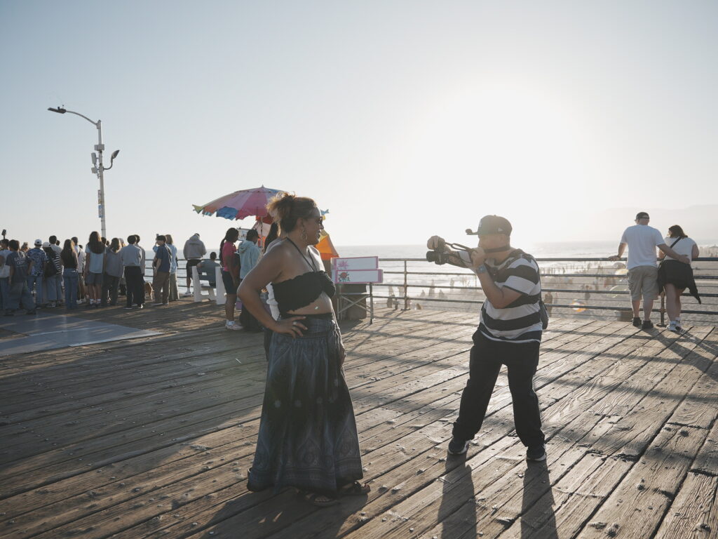 Photographer capturing a woman posing on Santa Monica Pier during golden hour, with the sun setting over the Pacific Ocean in the background.