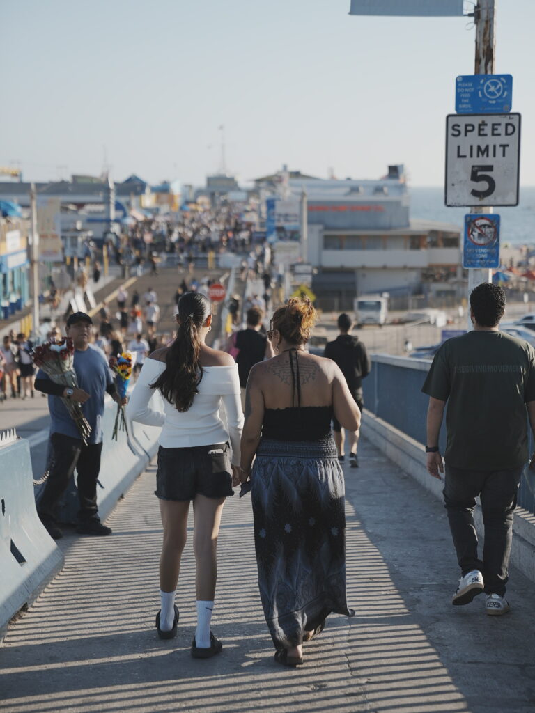 Mother and daughter walking hand in hand toward the bustling Santa Monica Pier, bathed in warm afternoon light with the ocean in the distance.