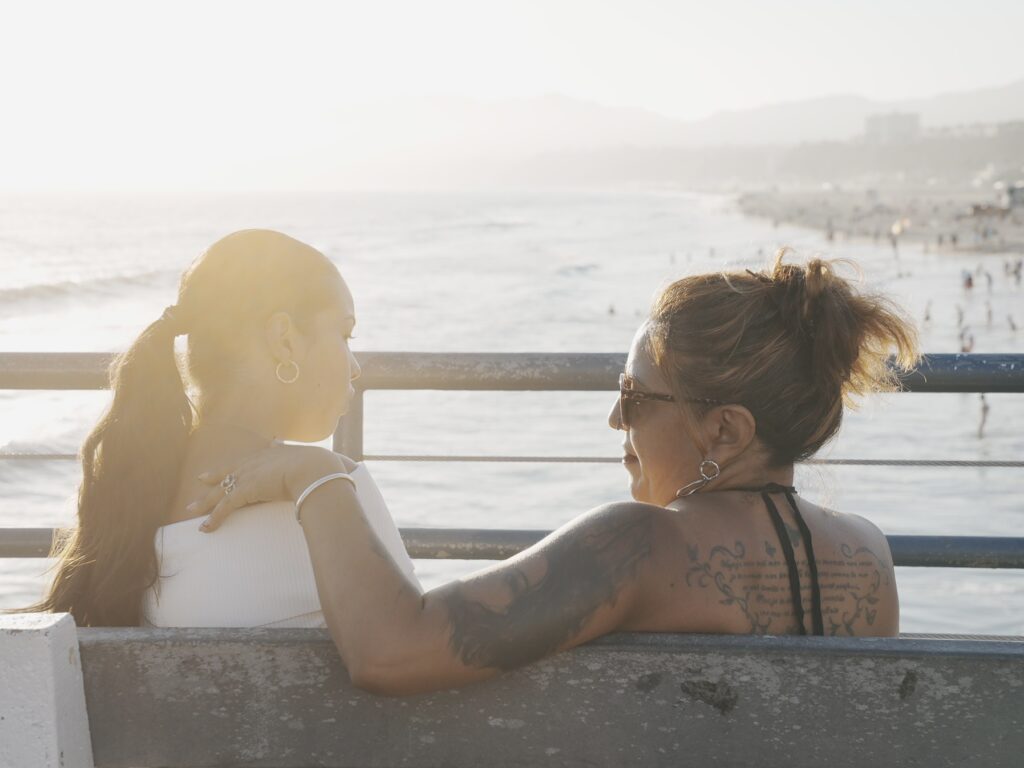 Mother and daughter sitting together on a bench at Santa Monica Pier, facing each other and talking as the sun sets over the Pacific Ocean.