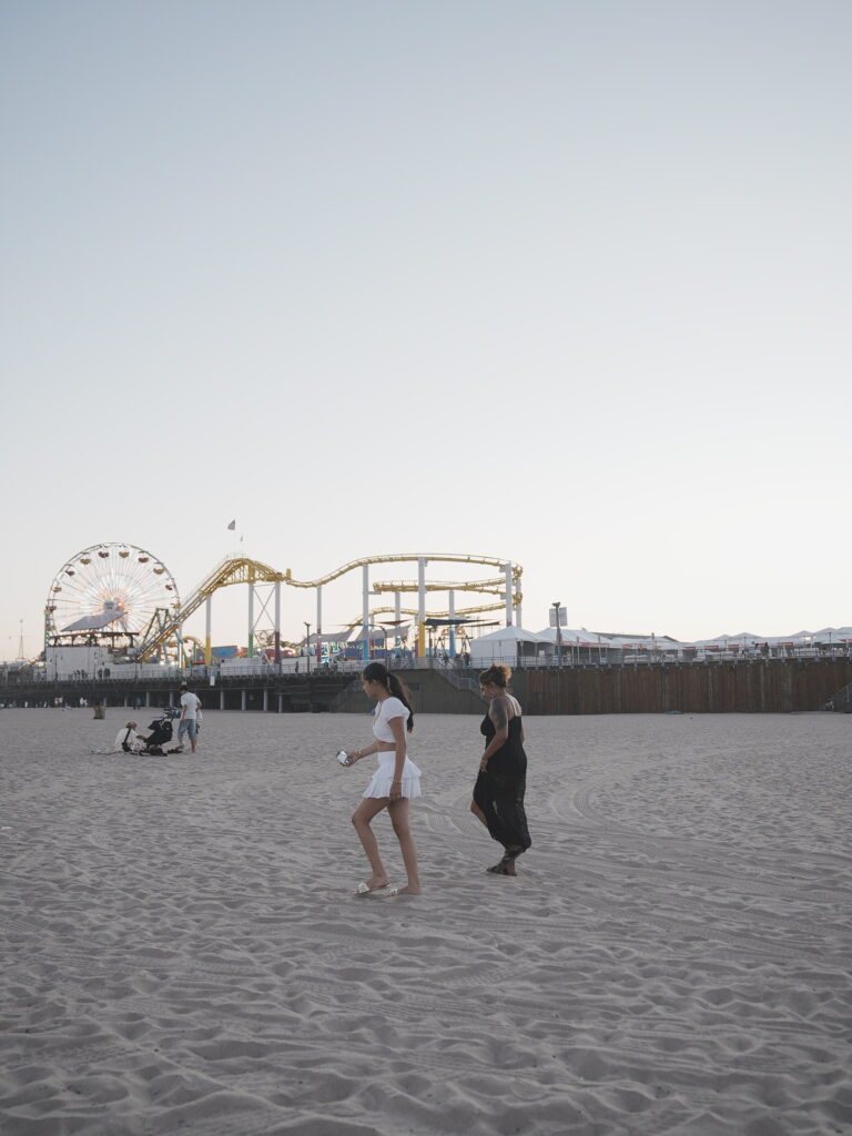 Mother and daughter walking barefoot on the sand near Santa Monica Pier at sunset, with the Ferris wheel and roller coaster in the background.