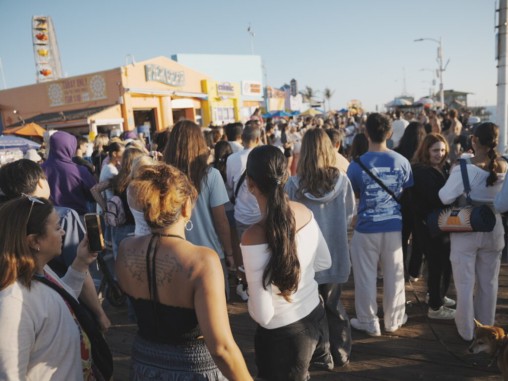 Crowds walking along the bustling Santa Monica Pier during golden hour, with colorful shops and the Pacific Park Ferris wheel in the background